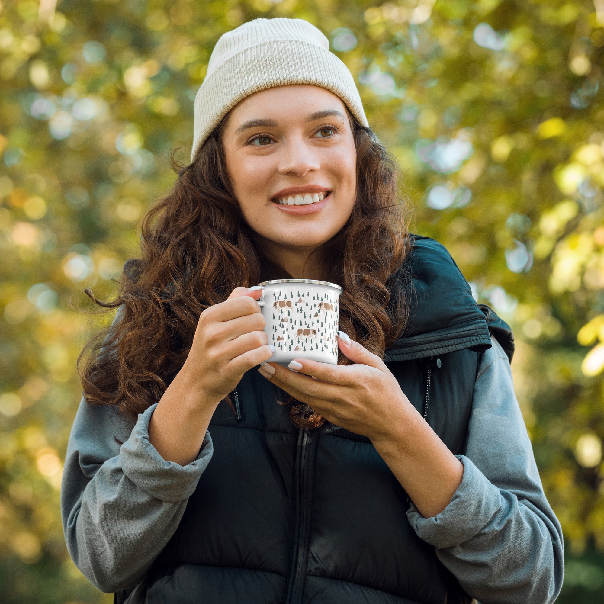 Woman holding lightweight 12 oz enamel camp mug. Features a modern pattern of Saint Bernard dogs, mountains and pine trees on a white background by Lucy and Norman