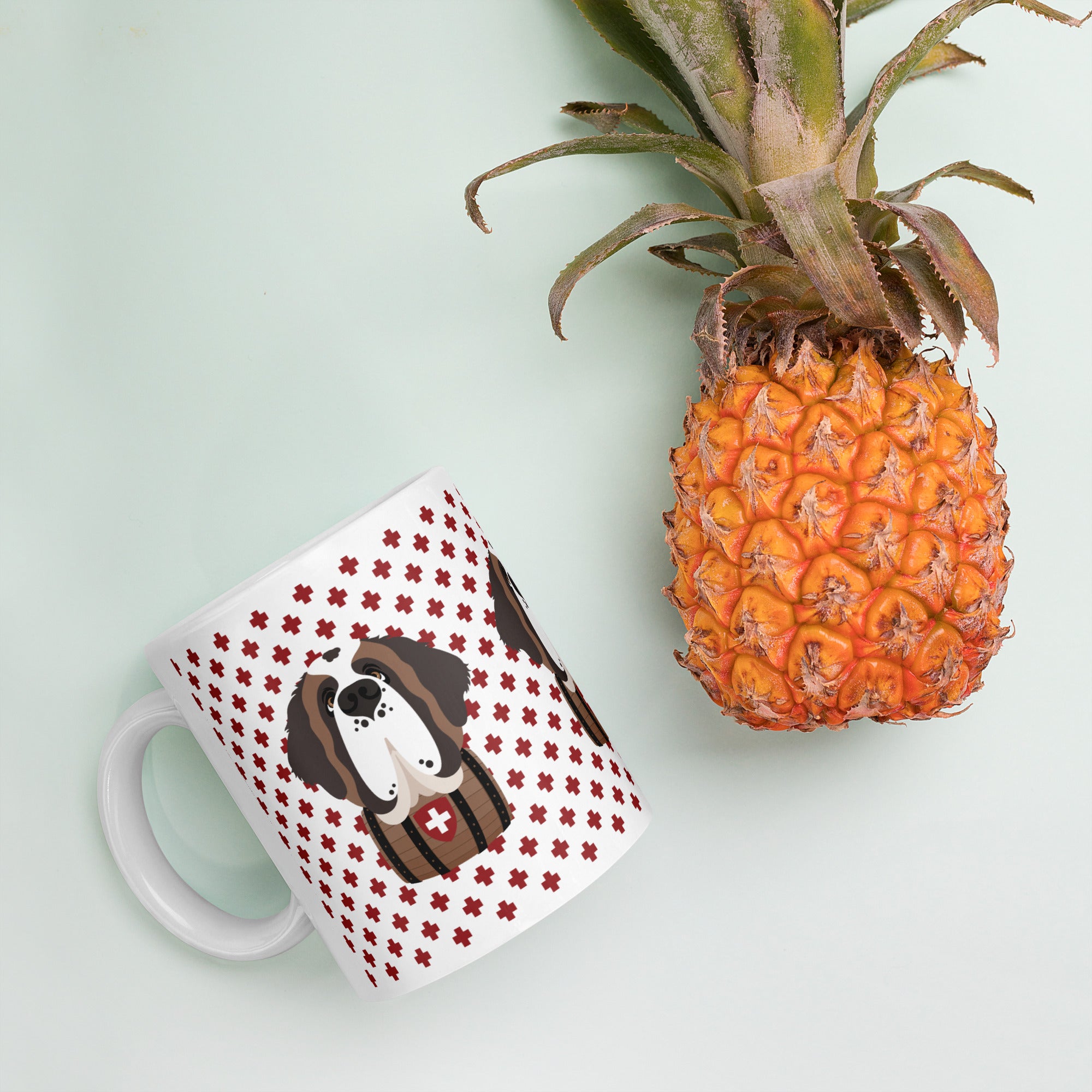 White ceramic 11 ounce mug featuring a cute illustrated Saint Bernard dog wearing a rescue barrel, surrounded by a red Swiss cross pattern. Perfect gift for St Bernard dog lovers and Swiss Alps fans by Lucy and Norman.