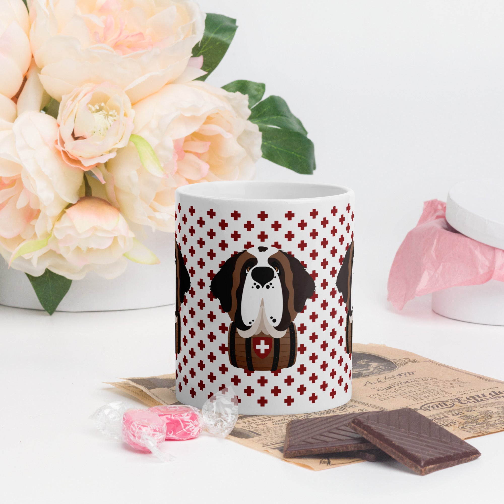 White ceramic mug featuring a cute illustrated Saint Bernard dog wearing a rescue barrel, surrounded by a red Swiss cross pattern. Perfect gift for St Bernard dog lovers and Swiss Alps fans by Lucy and Norman.