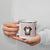 White enamel 12 ounce camp mug with silver rim featuring a cute illustrated Saint Bernard dog wearing a rescue barrel, surrounded by a red Swiss cross pattern. Perfect gift for St Bernard dog lovers and Swiss Alps fans by Lucy and Norman.