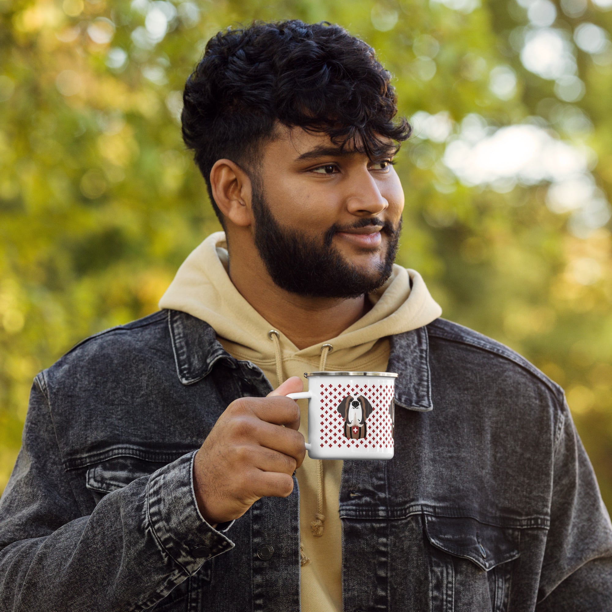 Man holding white enamel 12 ounce camp mug with silver rim featuring a cute illustrated Saint Bernard dog wearing a rescue barrel, surrounded by a red Swiss cross pattern. Perfect gift for St Bernard dog lovers and Swiss Alps fans by Lucy and Norman.