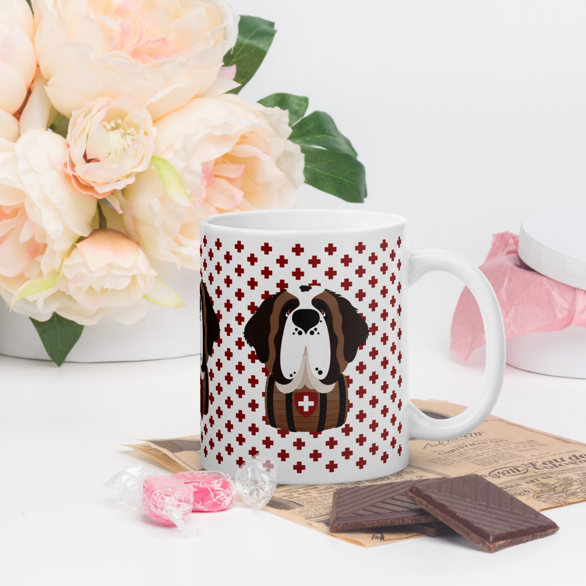 White ceramic 15 ounce mug featuring a cute illustrated Saint Bernard dog wearing a rescue barrel, surrounded by a red Swiss cross pattern. Perfect gift for St Bernard dog lovers and Swiss Alps fans by Lucy and Norman.